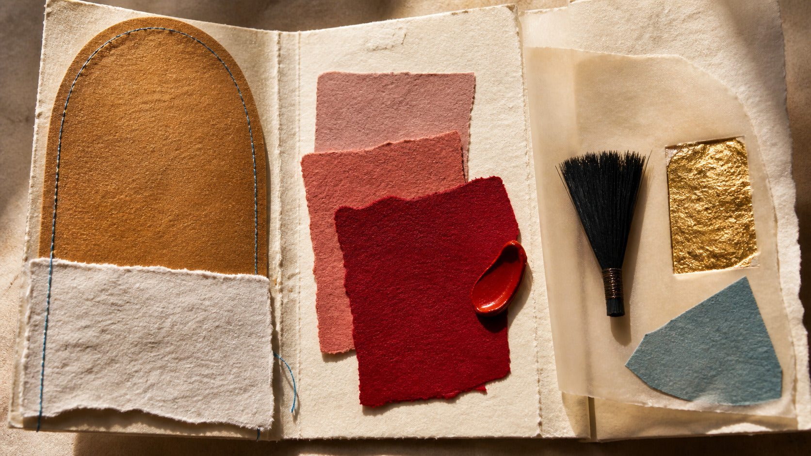 Open handmade-paper folio with cream and ochre layers, red colour swatches, black fibres, gold paper and blue stitching